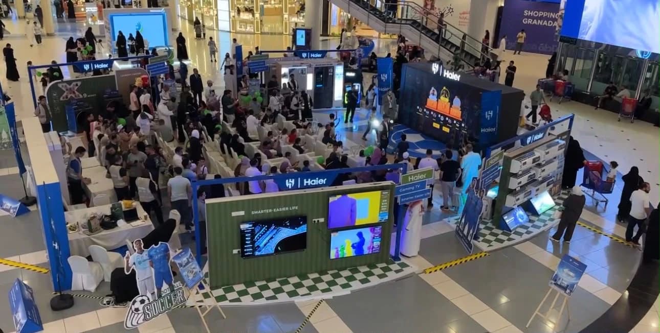 فعالية ترويجية في بهو مركز تجاري من الأعلى، فيها جمهور جالس ومسرح وشاشات ومنصات لعرض المنتجات
